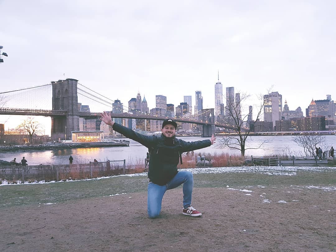 A photograph of Manuel kneeling on one knee and 
                    arms extended with a wide smile on his face. To his back, Brooklyn Bridge and New York's skyline.