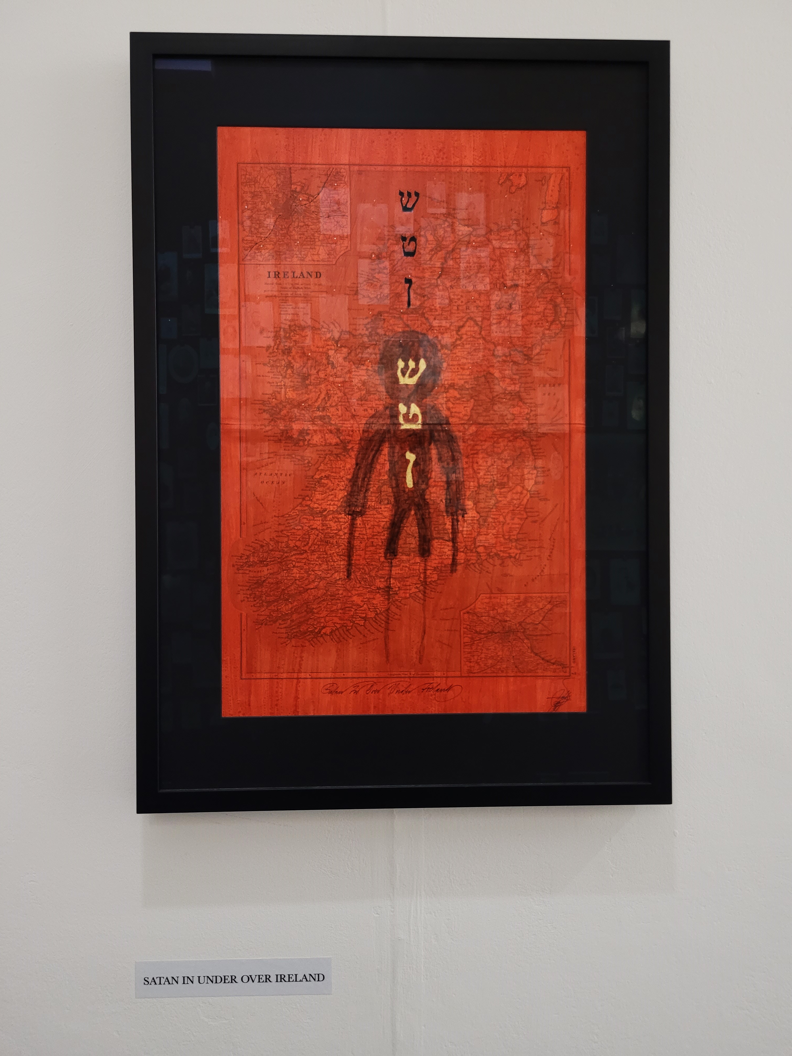 A photograph of one of the exhibition pieces: a map of Ireland painted in red and 
                            a black man figure over it. The description below the piece reads: Satan in under over Ireland