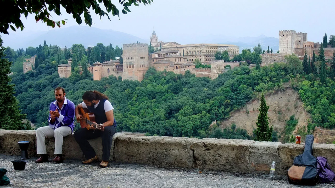 Photograph of The Alhambra from a viewpoint (Mirador de San Nicolás). Both a guitarist playing the guitar and a singer are sitting at the edge of the viewpoing.