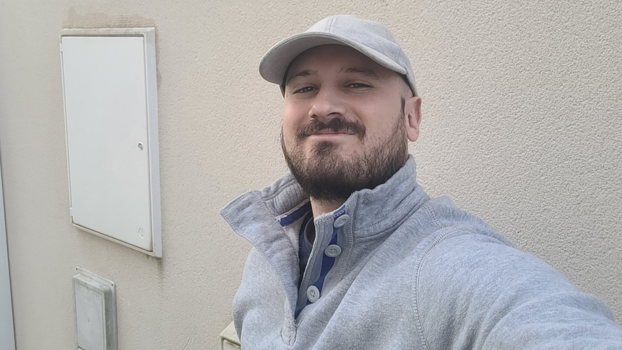 Selfie of Manuel wearing a grey cap and a grey jumper. No distinct background, just a wall.